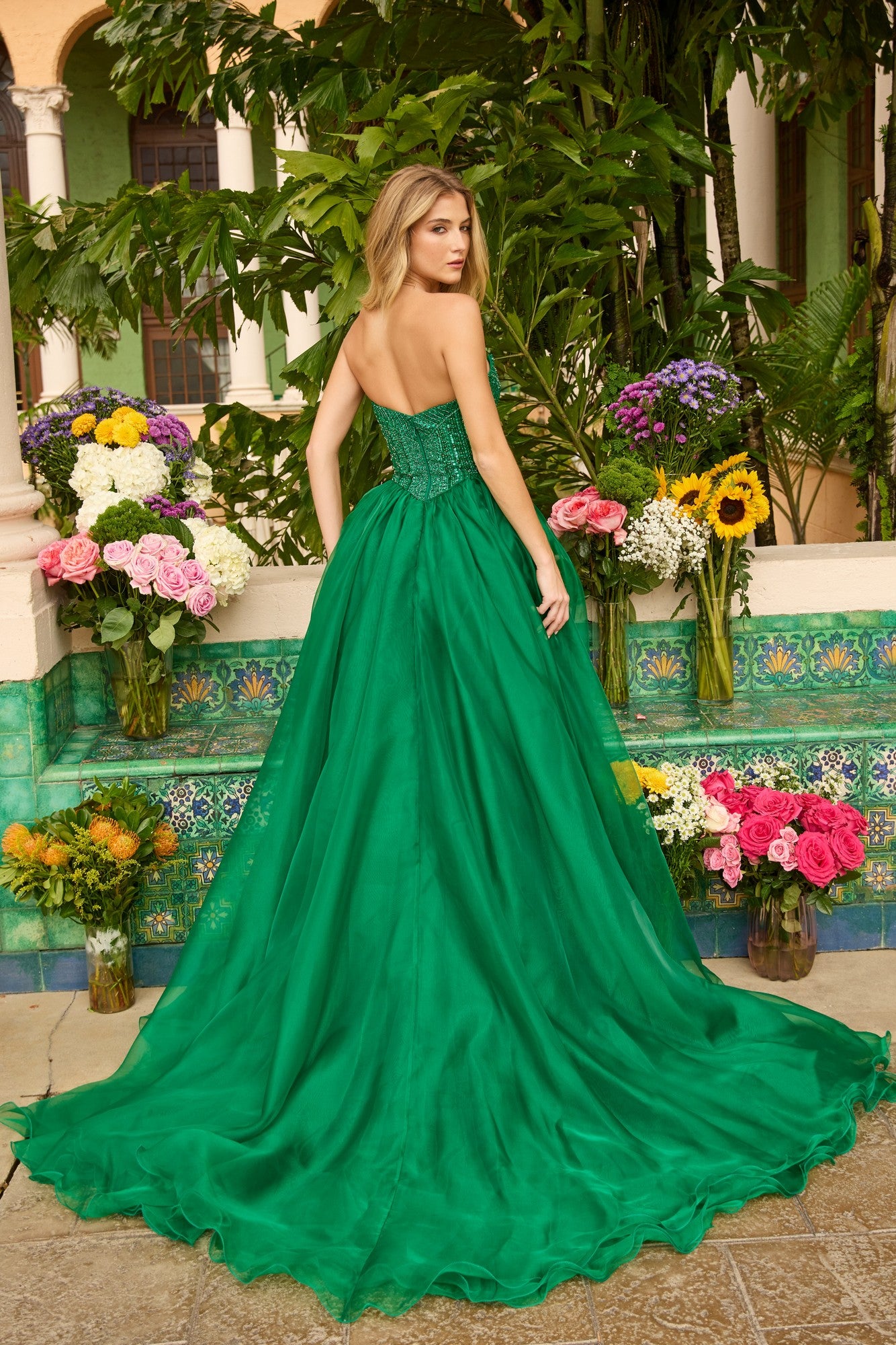 A woman stands outside among lush plants and vibrant flowers, her back to the camera, wearing the Long Prom Dress: Ava Presley 47255 featuring a strapless design and an embellished bodice.